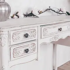 Un tavolo di legno, dipinto di bianco con un effetto anticato, con cassetti intagliati e pomelli decorativi.