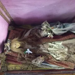 Bambola antica in una scarabattola, con capelli biondi, corona floreale e abiti sontuosi. Il suo significato è legato alla devozione e all'arte sacra.
