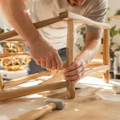 Uomo che ripara una sedia traballante, inserendo un tassello di legno. Martello e stoffa sul tavolo.
