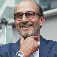 Uomo sorridente con occhiali, indossa un abito blu e un orologio Richard Mille, proprietario del brand.