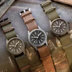 Tre orologi militari famosi Hamilton, con cinturini in pelle e tessuto, su sfondo metallico.