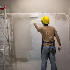 Operaio con casco giallo dipinge un muro grezzo con un rullo. Si può verniciare sopra l'impregnante, trasformando l'ambiente. Scala a pioli a sinistra.