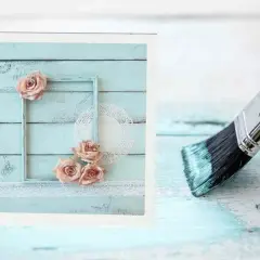 Creazione di una pittura shabby fai da te: un pennello stende vernice azzurra su assi di legno, mentre a sinistra un quadro decorato con rose e pizzo.