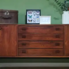 Mobile in legno scuro con cassetti e ante, decorato con una valigia vintage, quadri di farfalle e un vaso di fiori. Un esempio di restyling mobili che unisce passato e presente.