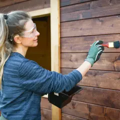 Donna con occhiali protettivi applica la finitura dopo impregnante su una parete di legno scuro.