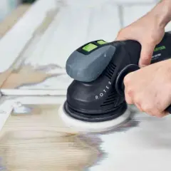 Mani che usano una levigatrice orbitale per rimuovere vecchia vernice e preparare il legno per una nuova finitura. Ecco come lucidare il legno per un risultato perfetto.