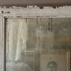 Dettagli di un vecchio specchio con cornice bianca scrostata, che riflette un ritratto e un drappo. Un esempio di come invecchiare uno specchio per un effetto anticato.