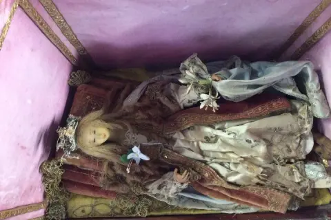Bambola antica in una scarabattola, con capelli biondi, corona floreale e abiti sontuosi. Il suo significato è legato alla devozione e all'arte sacra.