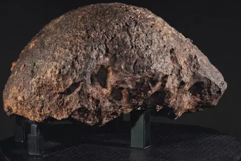 Massiccio meteorite, un esemplare di meteoriti rari, con una superficie ruvida e craterizzata, esposto su un piedistallo nero.