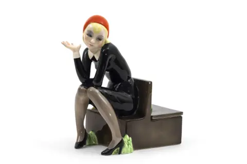 Figura in ceramica di donna con cappello rosso e abito nero, seduta su una panca. Un esempio per capire come riconoscere le bambole Lenci.
