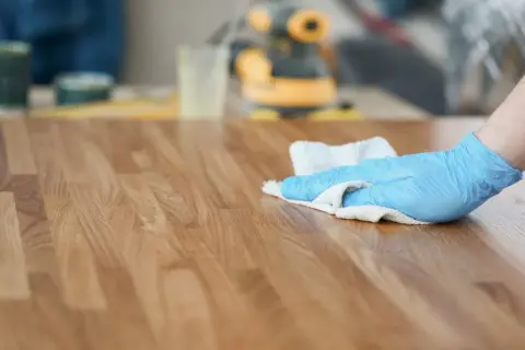 Mano con guanto blu che applica olio di lino per legno su una superficie in legno.