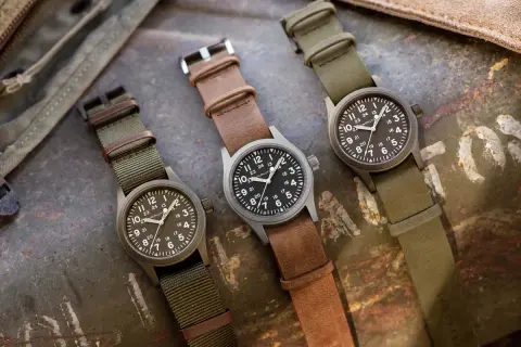 Tre orologi militari famosi Hamilton, con cinturini in pelle e tessuto, su sfondo metallico.