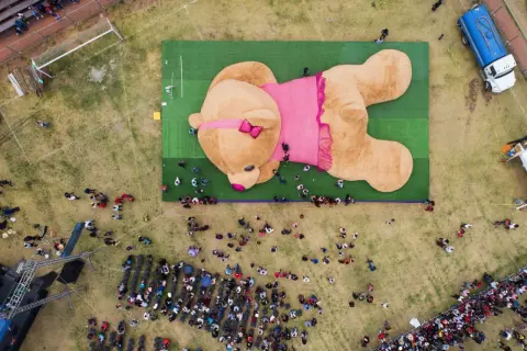 Un enorme orsacchiotto peluche, forse il peluche più costoso del mondo, giace su un prato verde, circondato da una folla curiosa.
