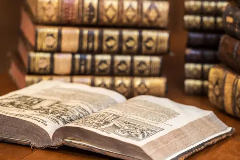 Un antico libro aperto mostra incisioni dettagliate, circondato da pile di volumi rilegati in pelle, testimoni di preziosi libri costosi.