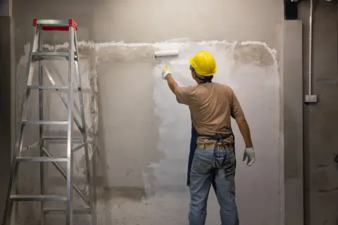 Operaio con casco giallo dipinge un muro grezzo con un rullo. Si può verniciare sopra l'impregnante, trasformando l'ambiente. Scala a pioli a sinistra.