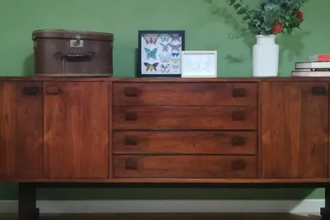 Mobile in legno scuro con cassetti e ante, decorato con una valigia vintage, quadri di farfalle e un vaso di fiori. Un esempio di restyling mobili che unisce passato e presente.