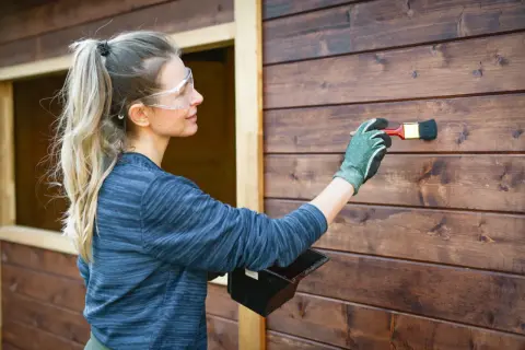Donna con occhiali protettivi applica la finitura dopo impregnante su una parete di legno scuro.