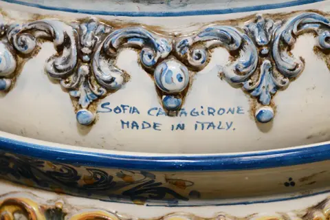 Dettaglio di un'opera in porcellana bianca e blu, con decorazioni barocche e la firma "Sofia Caltagirone Made in Italy".