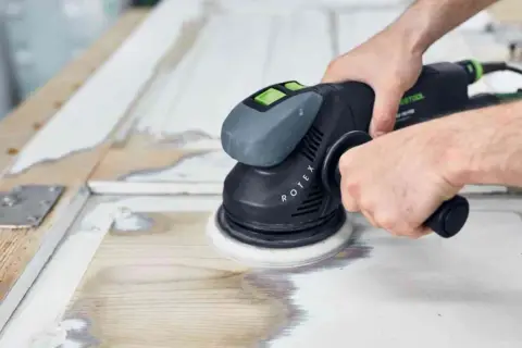 Mani che usano una levigatrice orbitale per rimuovere vecchia vernice e preparare il legno per una nuova finitura. Ecco come lucidare il legno per un risultato perfetto.