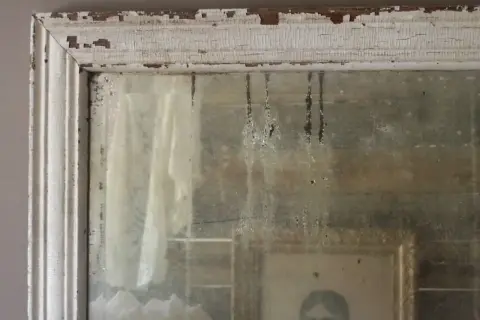 Dettagli di un vecchio specchio con cornice bianca scrostata, che riflette un ritratto e un drappo. Un esempio di come invecchiare uno specchio per un effetto anticato.