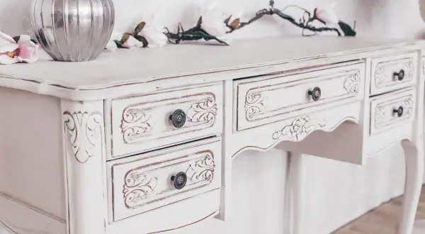 Un tavolo di legno, dipinto di bianco con un effetto anticato, con cassetti intagliati e pomelli decorativi.