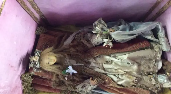 Bambola antica in una scarabattola, con capelli biondi, corona floreale e abiti sontuosi. Il suo significato è legato alla devozione e all'arte sacra.