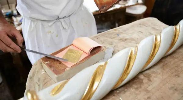 Artigiano applica foglia oro su un elemento decorativo bianco e oro. Scopri come si applica la foglia oro.