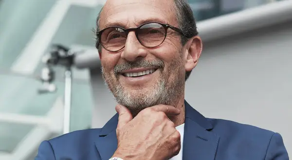 Uomo sorridente con occhiali, indossa un abito blu e un orologio Richard Mille, proprietario del brand.