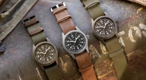 Tre orologi militari famosi Hamilton, con cinturini in pelle e tessuto, su sfondo metallico.