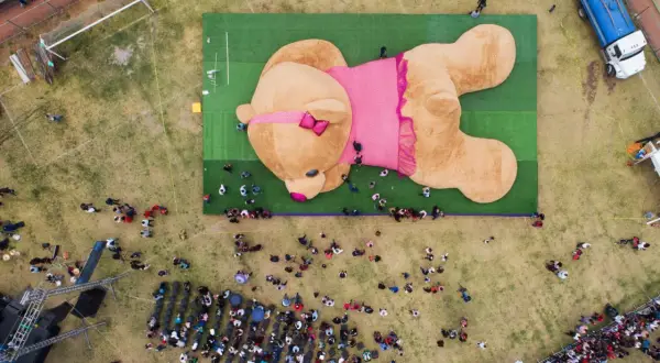 Un enorme orsacchiotto peluche, forse il peluche più costoso del mondo, giace su un prato verde, circondato da una folla curiosa.
