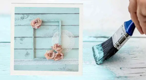 Creazione di una pittura shabby fai da te: un pennello stende vernice azzurra su assi di legno, mentre a sinistra un quadro decorato con rose e pizzo.