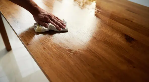 Mano che lucida un tavolo in legno con un panno, applicando una finitura a cera per un aspetto brillante e protetto.