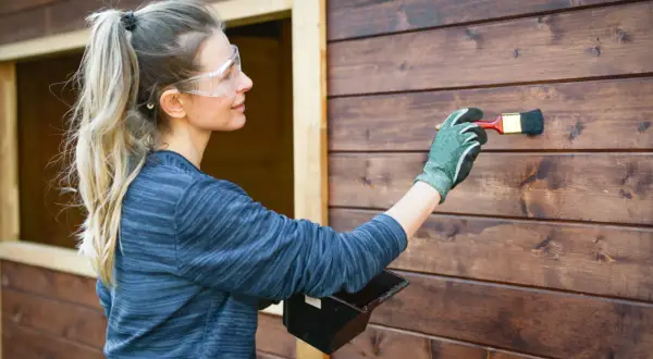 Donna con occhiali protettivi applica la finitura dopo impregnante su una parete di legno scuro.