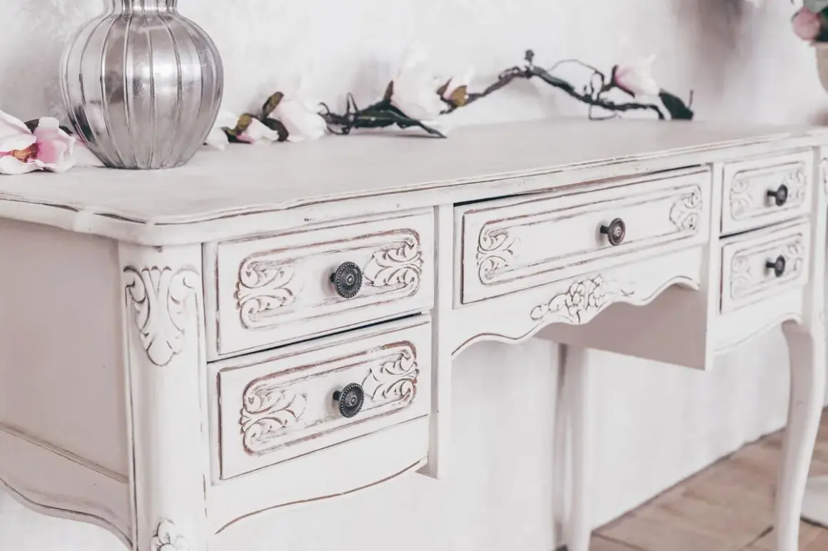 Un tavolo di legno, dipinto di bianco con un effetto anticato, con cassetti intagliati e pomelli decorativi.