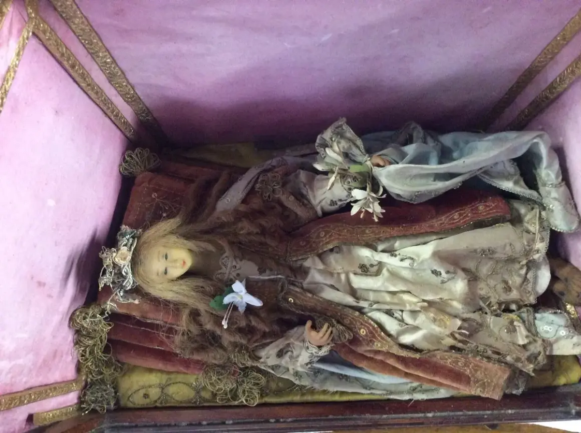 Bambola antica in una scarabattola, con capelli biondi, corona floreale e abiti sontuosi. Il suo significato è legato alla devozione e all'arte sacra.
