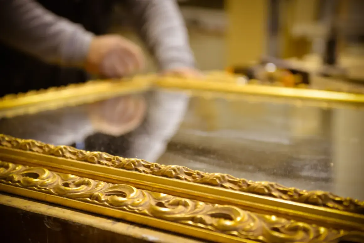 Mani esperte al lavoro per il restauro specchio antico. La cornice dorata e intarsiata riflette la cura del dettaglio.