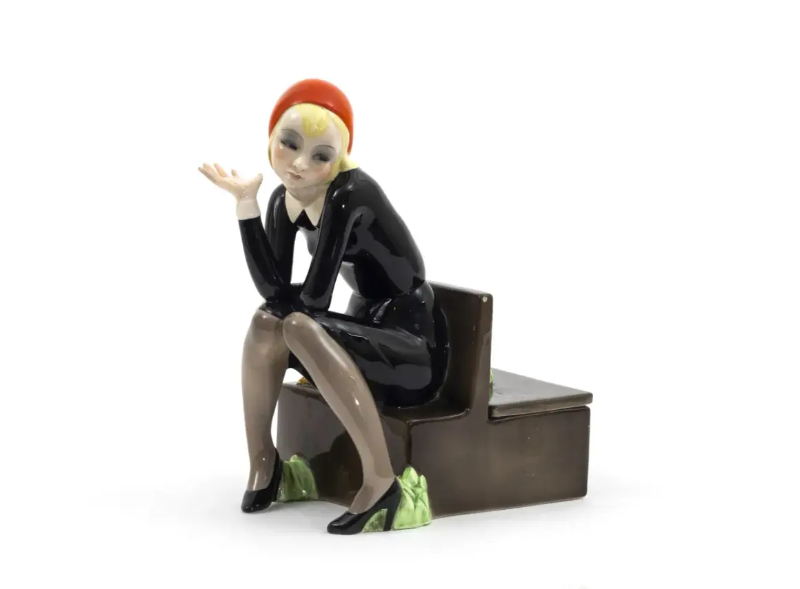 Figura in ceramica di donna con cappello rosso e abito nero, seduta su una panca. Un esempio per capire come riconoscere le bambole Lenci.