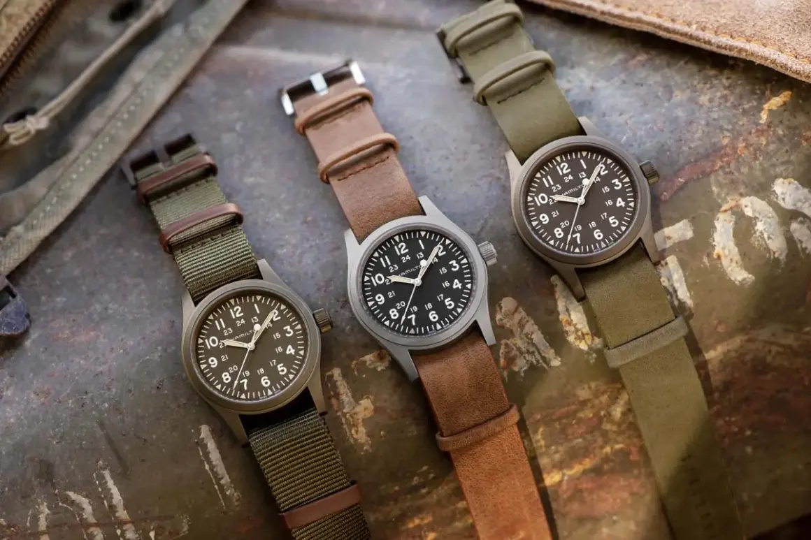 Tre orologi militari famosi Hamilton, con cinturini in pelle e tessuto, su sfondo metallico.