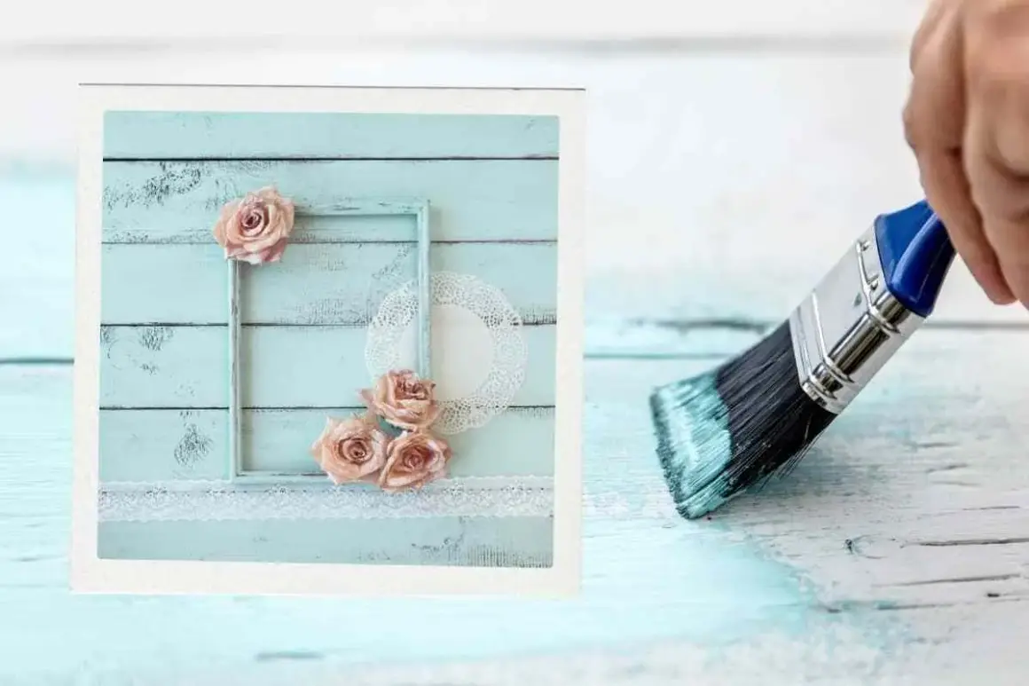 Creazione di una pittura shabby fai da te: un pennello stende vernice azzurra su assi di legno, mentre a sinistra un quadro decorato con rose e pizzo.