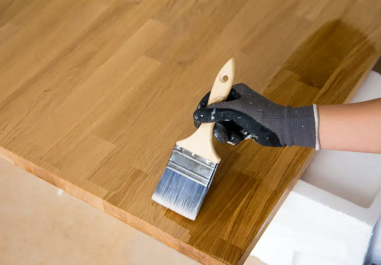 Mano guantata applica vernice su un piano in legno grezzo, completando il trattamento.