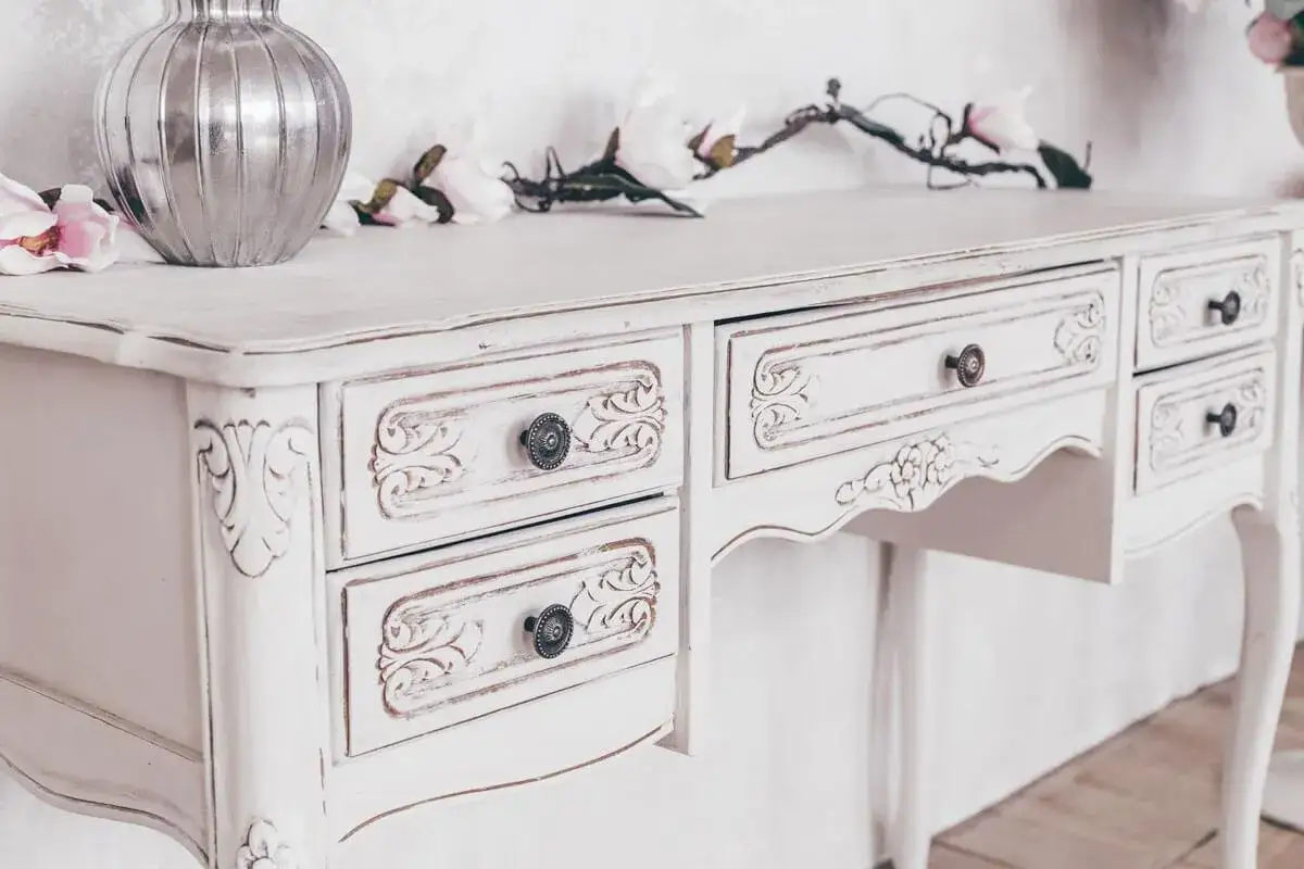 Un tavolo di legno dipinto di bianco, con cassetti intagliati e pomelli decorativi, abbellito da fiori e un vaso.