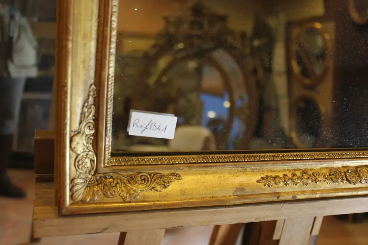 Specchio antico con cornice dorata e decorazioni floreali, in fase di restauro. Riflessi di altri specchi e arredi.