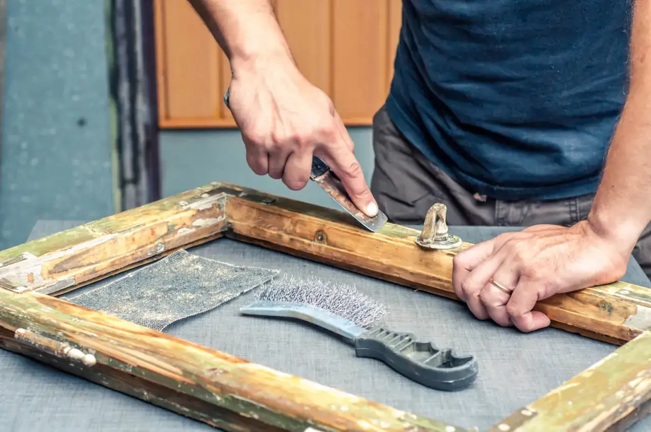 Mani che raschiano una vecchia cornice di legno, preparando la superficie per sverniciare con soda caustica. Strumenti da lavoro a portata di mano.
