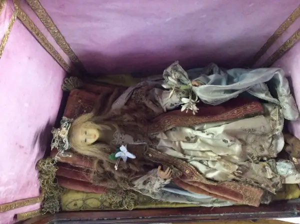 Bambola antica in una scarabattola, con capelli biondi, corona floreale e abiti sontuosi. Il suo significato è legato alla devozione e all'arte sacra.