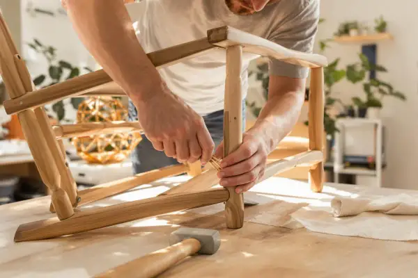 Uomo che ripara una sedia traballante, inserendo un tassello di legno. Martello e stoffa sul tavolo.
