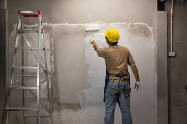 Operaio con casco giallo dipinge un muro grezzo con un rullo. Si può verniciare sopra l'impregnante, trasformando l'ambiente. Scala a pioli a sinistra.