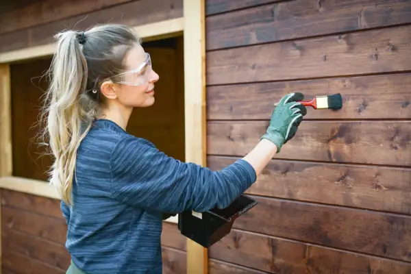 Donna con occhiali protettivi applica la finitura dopo impregnante su una parete di legno scuro.
