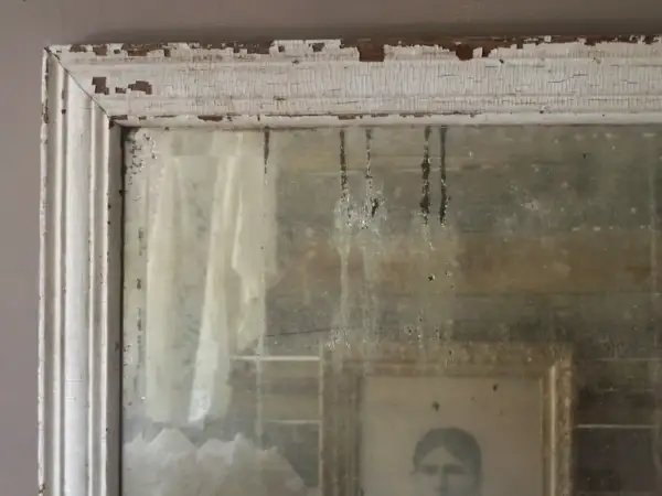 Dettagli di un vecchio specchio con cornice bianca scrostata, che riflette un ritratto e un drappo. Un esempio di come invecchiare uno specchio per un effetto anticato.